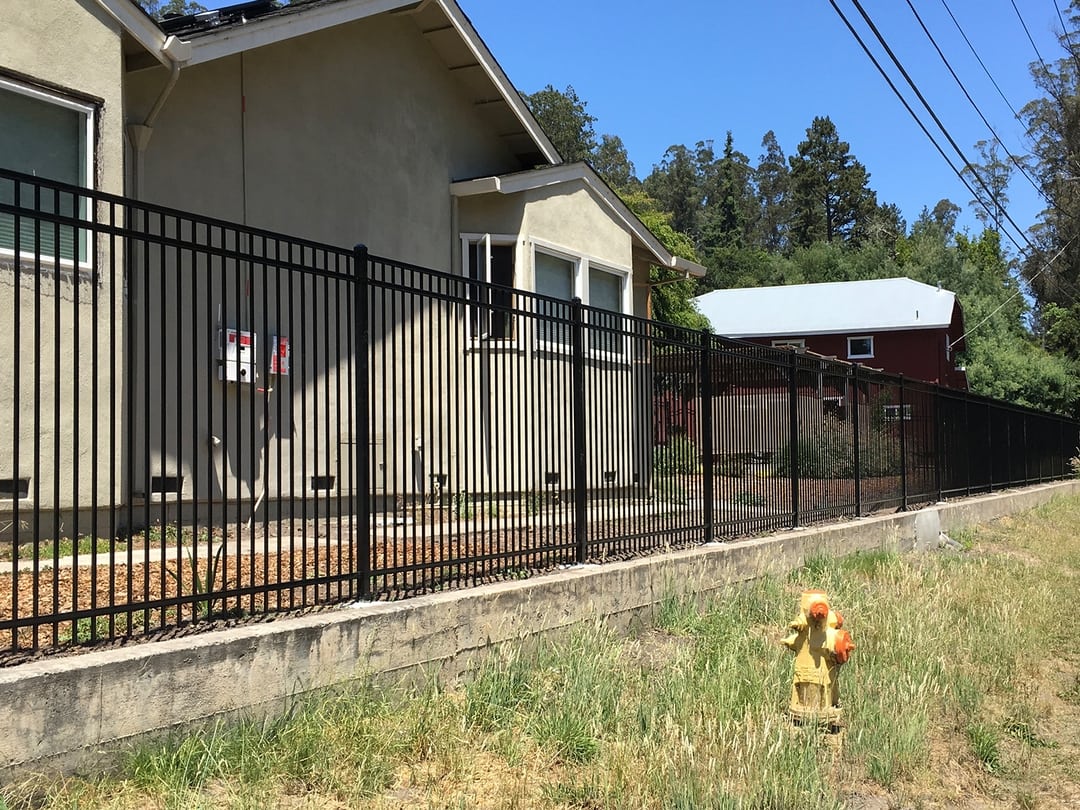 Flat top extruded, bottom, black fencing on a small concrete, retaining wall with a house behind