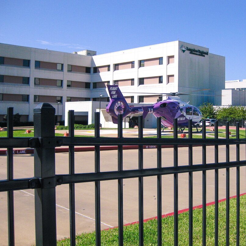 Extruded top, black fencing, surrounding a hospital facility with a  helicopter visible